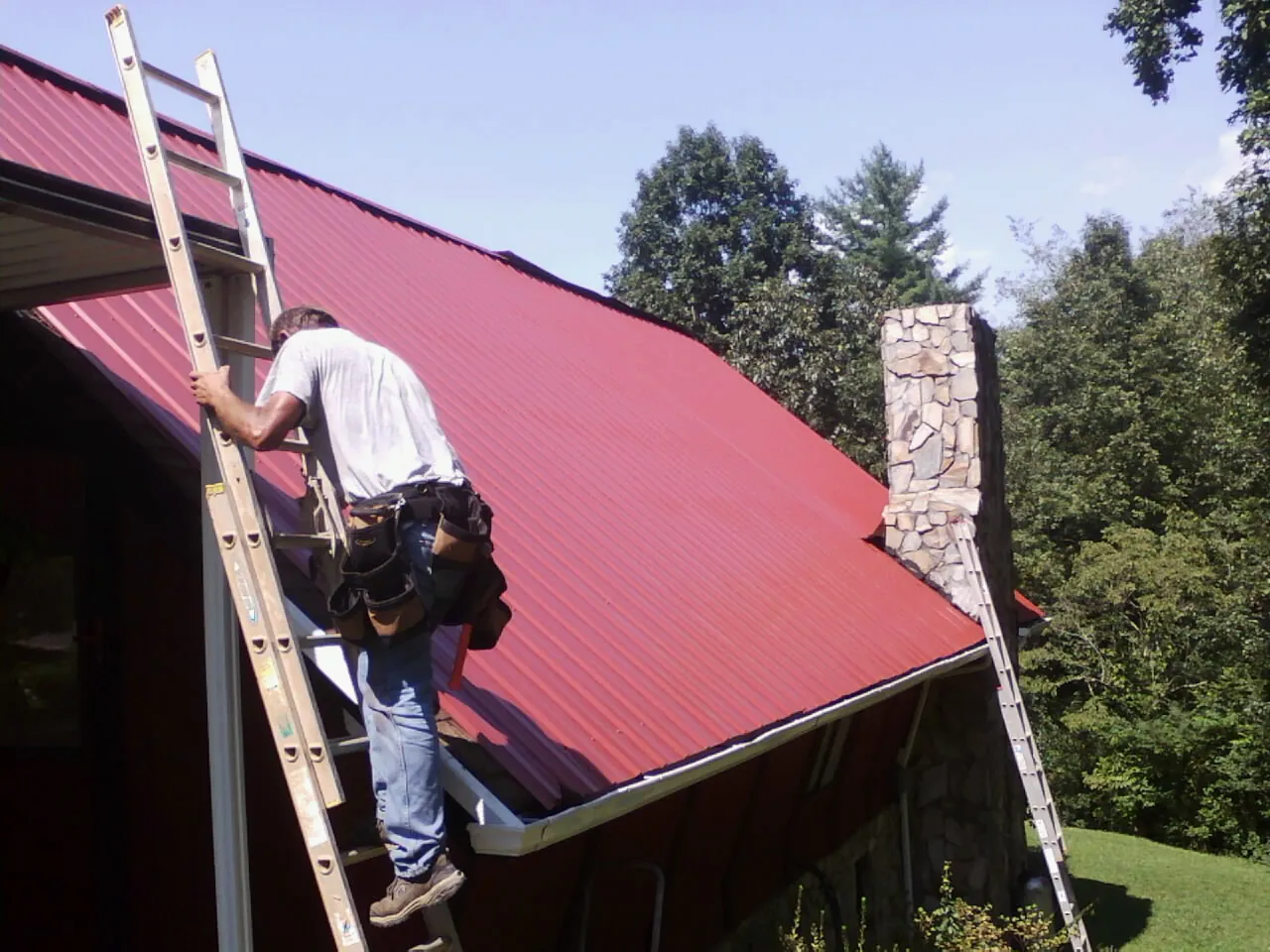 Roofer on a ladder beside a red metal roof and stone chimney — Soffit & Fascia Repair by Roofing Brothers in Powderly